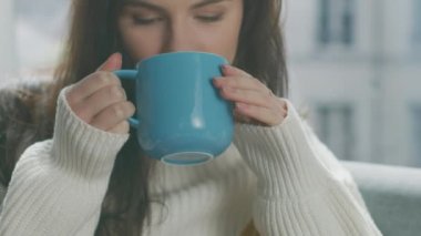 Genç Esmer Tutucu Kupa 'nın portresi Sıcak İçecek ve Zevkle İçmek. Beyaz örgü süveter giyen, rahat bir oturma odasında tek başına dinlenen tatlı kızın yakın çekimi.