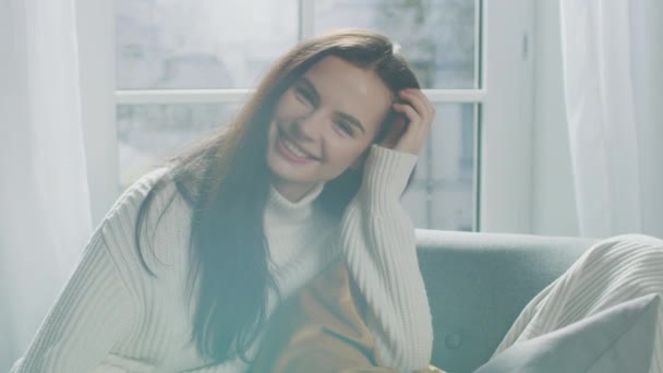 Retrato de bela morena jovem sorrindo encantadoramente enquanto olha para a câmera, escovando seu cabelo exuberante longe com um gesto flerte. Menina tímida vestindo camisola de malha branca rindo no quarto acolhedor — Vídeo de Stock