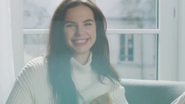 Portrait of Beautiful Young Brunette Smiling Charmingly while Looking at Camera, Brushing Her Lush Hair Away with a Flirtatious Gesture. Shy Girl Wearing White Knitted Sweater Laughing in Cozy Room — Stock Video