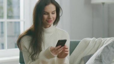 Akıllı Telefon Kullanan Genç Kadın, Sandalyede Otururken Çay İçen Kadın. Şehvetli Kız Süveter Giyiyor İnternet 'te, Sosyal medyada paylaşıyor, rahat bir dairede dinlenirken resimleri paylaşıyor
