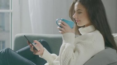 Akıllı Telefon Kullanan Güzel Genç Kadın, Sandalyede Otururken Çay İçen Kadın. Şehvetli Kız Süveter Giyiyor İnternet 'te, Sosyal medyada paylaşıyor, rahat bir dairede dinlenirken resimleri paylaşıyor