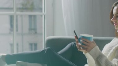 Akıllı Telefon Kullanan Güzel Genç Kadın, Sandalyede Otururken Çay İçen Kadın. Seksi Kız İnternet 'te Sörf Yapıyor, Sosyal medyada paylaşımlar yapıyor, resimlerini paylaşıyor. Yan Görünüm Yavaş Hareket