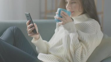 Akıllı Telefon Kullanan Güzel Genç Kadın, Sandalyede Otururken Çay İçen Kadın. Şehvetli Kız Süveter Giyiyor İnternet 'te, Sosyal medyada paylaşıyor, rahat bir dairede dinlenirken resimleri paylaşıyor