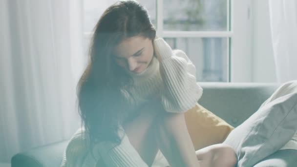 Retrato da jovem mulher sorrindo encantadoramente enquanto sentada em um sofá, escova seu cabelo exuberante longe com um gesto flerte. Menina tímida vestindo camisola de malha branca e shorts relaxa em um sofá — Vídeo de Stock