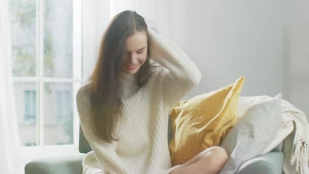 Krásná mladá žena s okouzlujícím úsměvem na kameru, zatímco sedí na židli. Roztomilé dívka s chaotické vlasy na sobě nadměrné bílé pletený svetr uvolní v neděli ráno v její útulný byt — Stock video