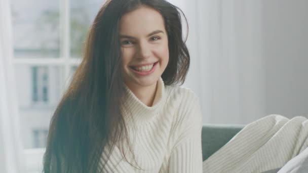 Jovem morena sorrindo encantadoramente para a câmera enquanto sentado na cadeira. Menina Sensual tímida com o cabelo bagunçado vestindo camisola de malha branca de grandes dimensões relaxa no domingo de manhã em seu aconchegante apartamento — Vídeo de Stock