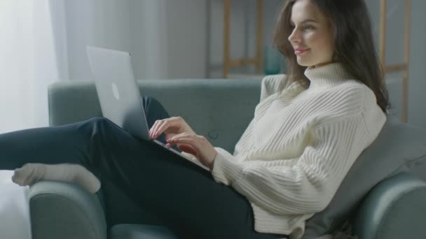 Piękna młoda kobieta pracuje na laptopie siedząc na krześle. Zmysłowa dziewczyna w swetrze działa na notebooku; Studia, Surfs Internet, Korzystanie z mediów społecznościowych podczas relaksu w przytulnym mieszkaniu — Wideo stockowe