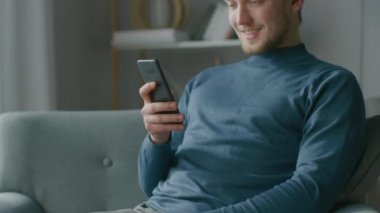 Oturma odasında sandalyesinde otururken akıllı telefon kullanan yakışıklı bir gencin portresi. Yaratıcı Freelancer Evde Gevşiyor, İnternette Sörf Yapıyor, Sosyal Medya Kullanıyor, Mesaj Gönderen Videolar İzliyor