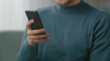 Oturma odasında sandalyesinde otururken akıllı telefon kullanan yakışıklı bir gencin portresi. Yaratıcı Freelancer Evde Gevşiyor, İnternette Sörf Yapıyor, Sosyal Medya Kullanıyor, Mesaj Gönderen Videolar İzliyor