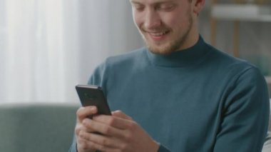 Oturma odasında sandalyesinde otururken akıllı telefon kullanan yakışıklı bir gencin portresi. Yaratıcı Freelancer Evde Gevşiyor, İnternette Sörf Yapıyor, Sosyal Medya Kullanıyor, Mesaj Gönderen Videolar İzliyor