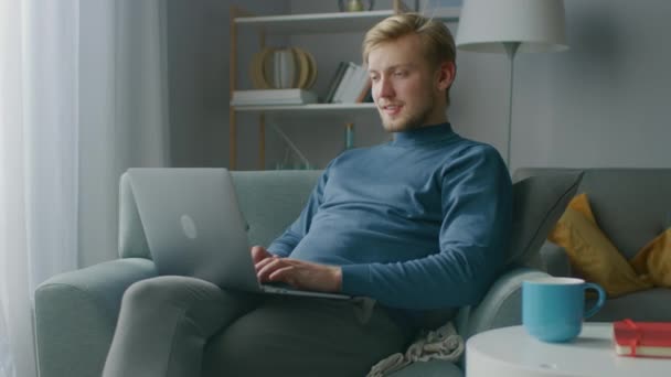 Πορτρέτο του Όμορφου Ξανθού Νέου που εργάζεται σε έναν φορητό υπολογιστή, ενώ κάθεται σε μια καρέκλα στο άνετο σαλόνι του. Creative Freelancer Relaxes at Home, Surfs Internet, Χρησιμοποιεί τα Social Media και Relaxes — Αρχείο Βίντεο