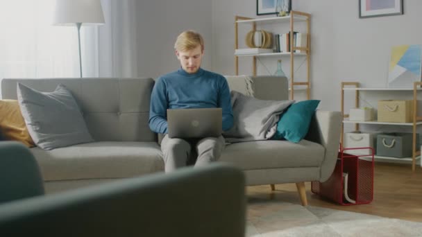 Retrato del joven guapo y rubio que trabaja en una computadora portátil, mientras está sentado en un sofá en su acogedora sala de estar. Freelancer creativo se relaja en el hogar, navega por Internet, utiliza las redes sociales y se relaja — Vídeo de stock