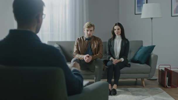 Молодая пара на сеансе психотерапевта. Обратный взгляд терапевта: молодые люди сидят на диване-аналитике, обсуждают психологические травмы, проблемы отношений и страдания — стоковое видео