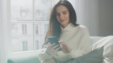 Akıllı Telefonu Mutlulukla Kullanan Güzel Genç Kadın, Sandalyede Otururken. Örgü ören, internette sörf yapan, sosyal medyada paylaşan, rahat bir dairede dinlenirken resim paylaşan seksi kız.
