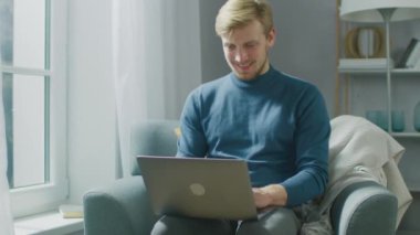 Yakışıklı Sarışın Genç Adam 'ın portresi. Oturma odasında bir sandalyede otururken dizüstü bilgisayarda çalışıyor. Yaratıcı Freelancer Evde Gevşiyor, İnternette Sörf Yapıyor, Sosyal Medya Kullanıyor ve Rahatlıyor