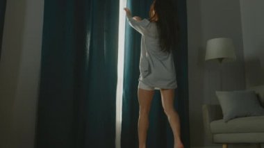 Güzel Genç Esmer Perdeleri açıyor Sabah Güneşinin rahat dairesini aydınlatmasına izin vermek için, Odada güzelce ve profesyonelce dans etmeye devam ediyor. Yavaş Hareket Düşük Açı Kenar Görünümü