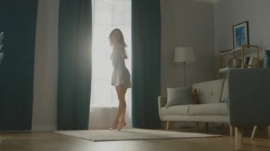 Güzel Genç Esmer Perdeleri açıyor Sabah Güneşinin rahat dairesini aydınlatmasına izin vermek için, Odada güzelce ve profesyonelce dans etmeye devam ediyor. Yavaş Hareket Düşük Açı Kenar Görünümü