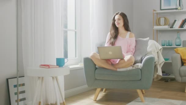 Retrato de bela jovem morena funciona no computador portátil enquanto bebe chá em seu aconchegante apartamento. Linda menina doce vestindo camisola rosa, passa o tempo navegando na Internet, usando as mídias sociais — Vídeo de Stock