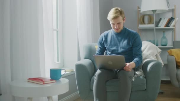 Портрет красивого молодого человека, работающего за ноутбуком, сидя на стуле в его уютном, счастливо празднует свой успех, поднимая руки в праздничный жест — стоковое видео