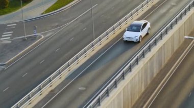 Bulutlu Bir Gün sırasında Şehir Kavşağında Modern Lüks Beyaz Elektrikli Geçit Sürüşünün Hava Aracı Görünümü. Otoyoldaki bir köprüde pilli araba kullanılıyor. Panoramik Çatılı Geleceğin SUV 'si.
