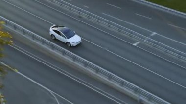 Bulutlu Bir Gün sırasında Şehir Yolu 'nda Modern Lüks Beyaz Elektrikli Geçit Sürüşünün Hava Aracı Görünümü. Pille çalışan araba otobandaki köprüden çıkıyor. Panoramik Çatılı Geleceğin SUV 'si.