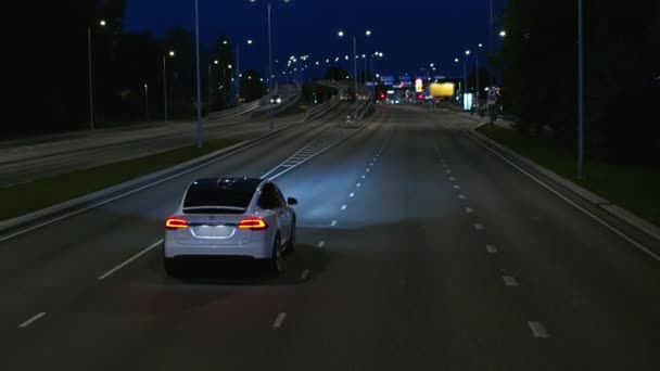 LuchtDrone Volg Shot van een moderne luxe witte elektrische SUV rijden op de stedelijke snelweg. Batterij aangedreven auto bewegen door lege weg met koplampen 's nachts. Futuristische Autonome Zelfrijdende Auto. — Stockvideo