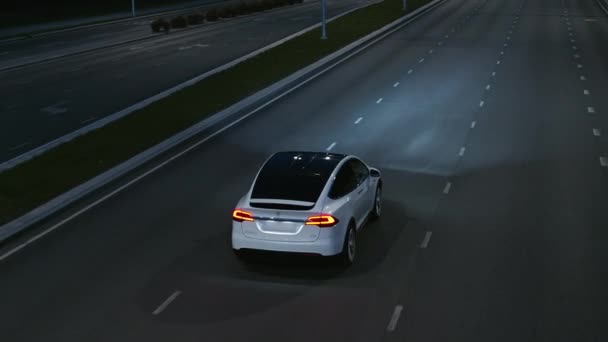 Drohne aus der Luft fotografiert ein modernes weißes Elektro-SUV, das auf der Stadtautobahn unterwegs ist. Batteriebetriebenes Auto, das nachts mit Scheinwerfern durch leere Straßen fährt. Futuristisches autonom fahrendes Auto.