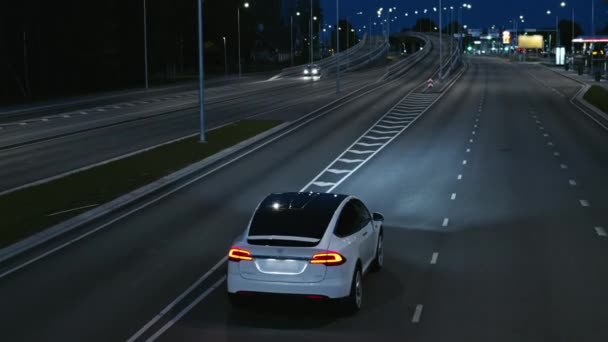 Εναέρια Drone Follow Shot of a Modern Luxury White Electric SUV Οδήγηση στην αστική εθνική οδό τη νύχτα. Battery Powered Car Ενεργοποιεί Σήμα στροφής και αλλαγές Lane. Φουτουριστικό αυτόνομο αυτοκινούμενο αυτοκίνητο. — Αρχείο Βίντεο