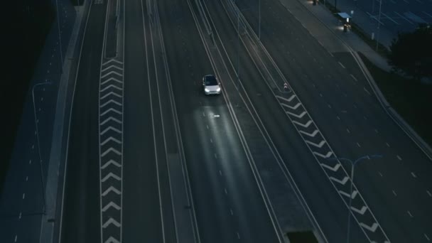 Vista aérea del drone de un SUV eléctrico blanco de lujo moderno que conduce en la carretera urbana por la noche. El coche alimentado por batería activa la señal de giro y cambia de carril. Coche futurista con faros de xenón . — Vídeos de Stock