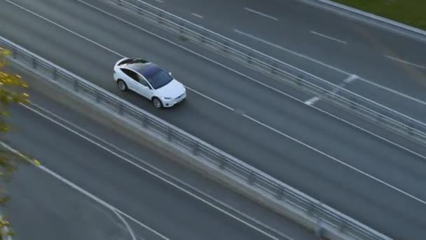 Vista aérea do drone de um cruzamento elétrico branco luxuoso moderno que conduz na estrada urbana durante um dia nublado. Carro alimentado por bateria sai de uma ponte em uma estrada. SUV futurista com telhado panorâmico . — Vídeo de Stock