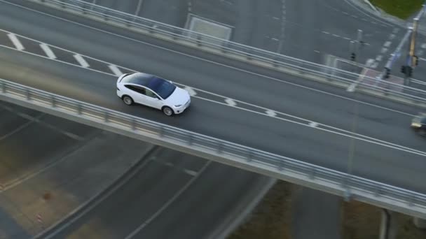 Vista aérea del dron de un cruce eléctrico blanco de lujo moderno que conduce por la carretera urbana durante un día nublado. El coche alimentado por batería conduce sobre un puente en una carretera. SUV futurista con techo panorámico . — Vídeos de Stock