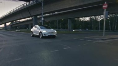 Akşam vakti Şehir Yolunda Modern Lüks Beyaz Elektrikli Geçit Sürüşü. Pille çalışan araba trafik ışıklarıyla kavşağa giriyor. Panoramik Çatı ve Xenon Işıklı Gelecekçi SUV.