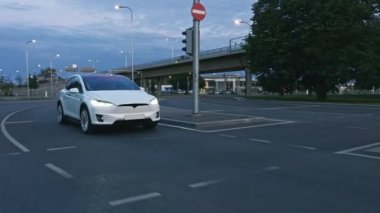 Akşam vakti Şehir Yolunda Modern Lüks Beyaz Elektrikli Geçit Sürüşü. Pille çalışan araba trafik ışıklarıyla kavşağa giriyor. Panoramik Çatı ve Xenon Işıklı Gelecekçi SUV.