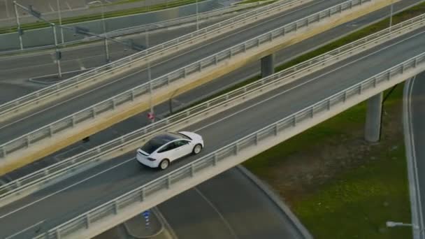 Vista aérea del dron de un cruce eléctrico blanco de lujo moderno conduciendo por Urban Road durante un día soleado. El coche alimentado por batería conduce sobre un puente en una carretera. Futurista autónomo auto conducción SUV . — Vídeo de stock