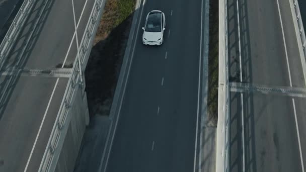 Slow Motion LuchtDrone View of a Modern Luxury White Electric SUV Driving on Highway. Batterij aangedreven auto bewegen door lege weg tussen viaducten. Futuristische Crossover met panoramisch dak. — Stockvideo