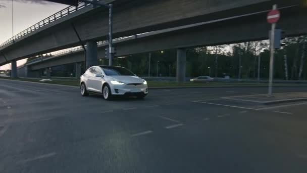 Moderner weißer Elektro-Crossover, der abends auf der Stadtstraße unterwegs ist. Batteriebetriebenes Auto passiert Kreuzung mit Ampel Futuristisches SUV mit Panoramadach und Xenonlicht. — Stockvideo