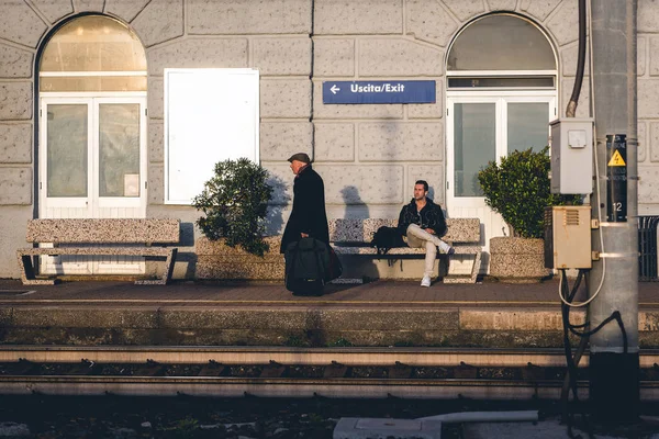 Senior man is leaving train station while young man is sitting on a bench near station platform. — Stock Photo, Image