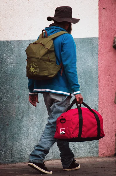 Poor man with a rucksack and red bag is walking along the street. — Stock Photo, Image