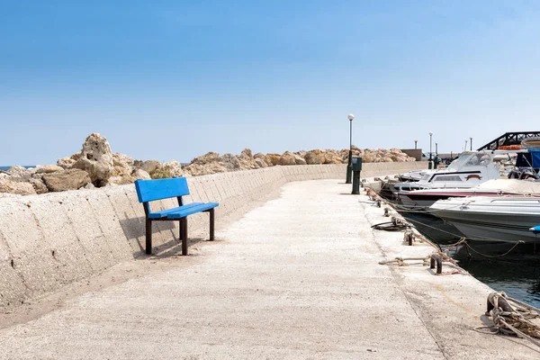 Pequeño paseo marítimo con banco azul en el pequeño puerto pesquero de la ciudad de Faliraki en la isla de Rodas, Grecia . — Foto de Stock