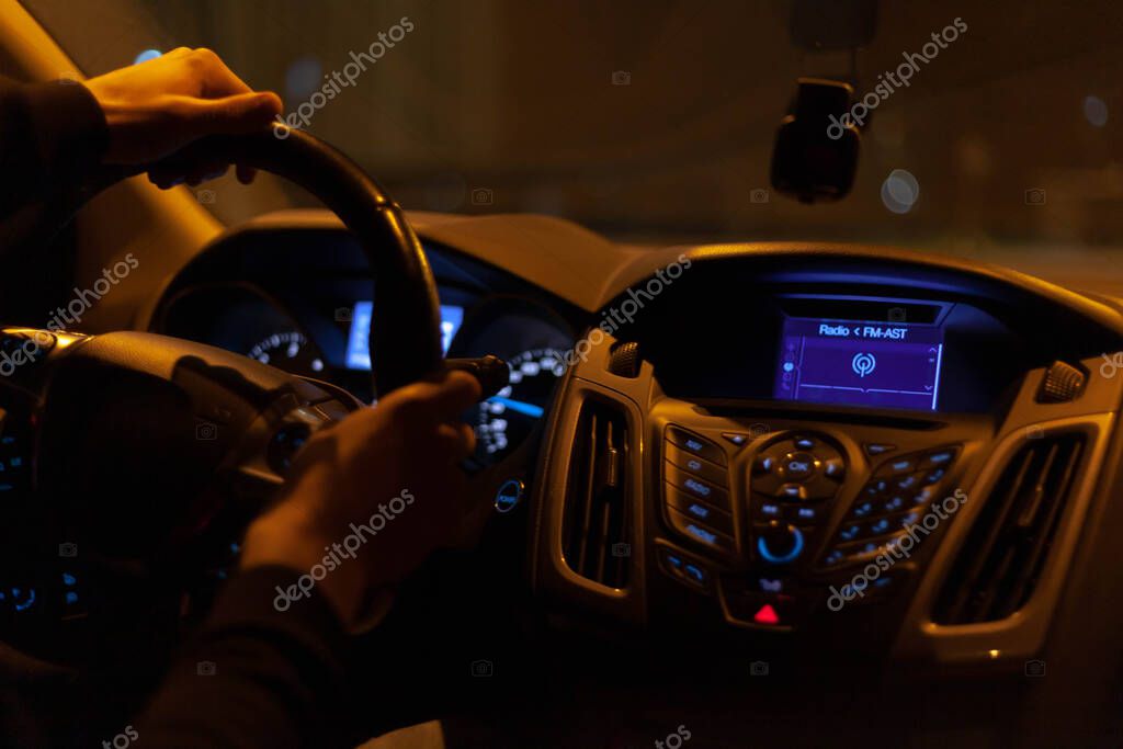A late night ride, dark moody car interior with lonely driver hands on wheel taking a sharp turn right, yellow street lighting. Traditional radio fm-ast symbol on the dashboard, view from behind