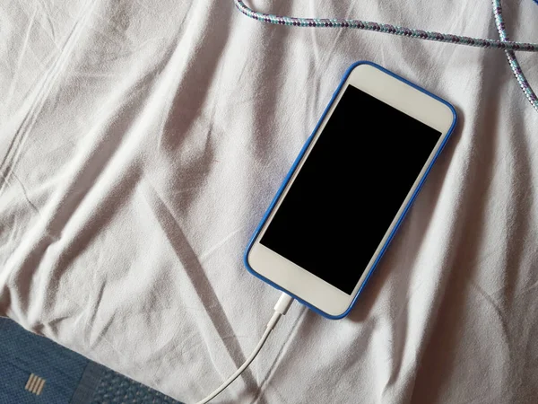 A white no name phone charging on a messy bed with creased bedsheet composition. Waking up in the morning with your smartphone or overnight battery charging concept.