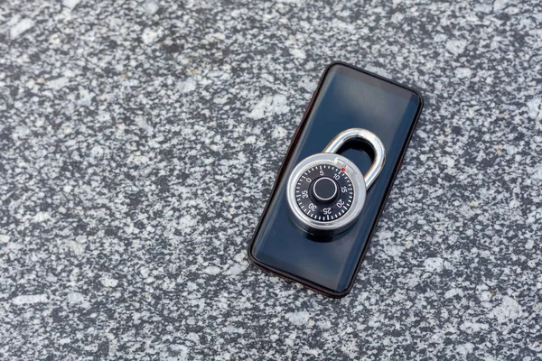 A modern black no name smartphone and a shiny round simple combination padlock laying on top of it, number code lock as a mobile device security symbol concept on a granite background, copyspace