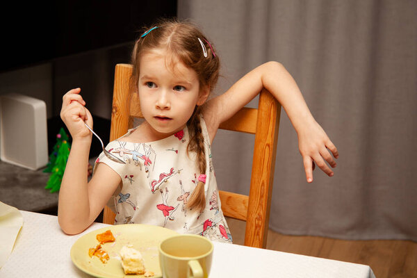 Little young girl eating her food holding a fork in her hand sitting on a chair in a sloppy, careless relaxed pose / manner. Arm on the chair back. Fussy eater, teaching table manners abstract concept