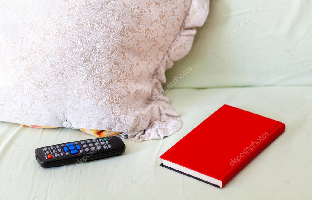 TV remote, red book and a pillow set of objects laying on an unmade bed / sofa Leisure, free time concept, staying home indoor activities, boredom, things to do Book vs tv alternative abstract, nobody