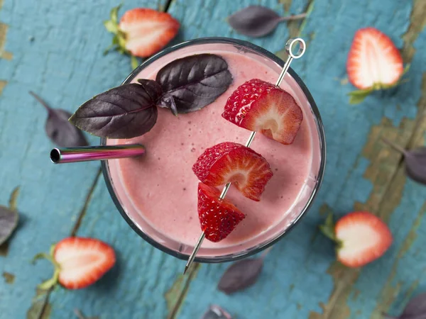 Top view on the strawberry smoothie in a glass with metal straw, basil leaves, strawberries on cocktail stick on old wooden surface. Halves of strawberries on a turquoise surface