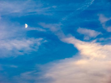 Parlak mavi bulutlu bir gökyüzünde yarım ay Gün batımı parıltısı ve soluk renk tonuyla