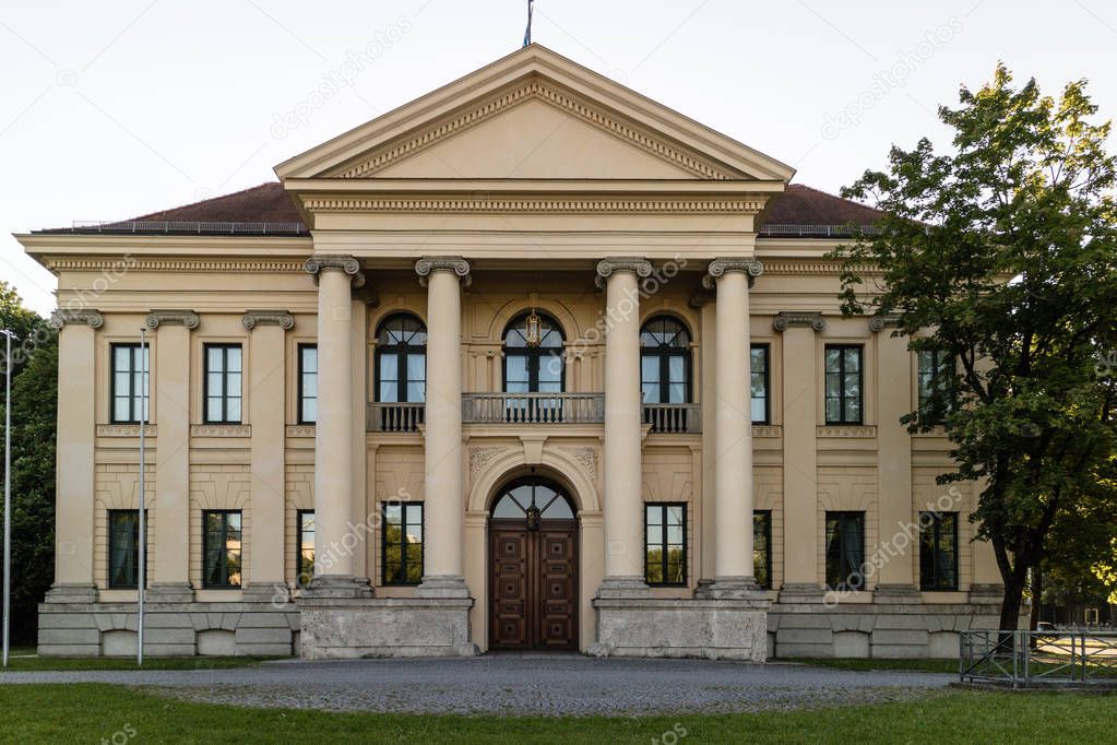 Historic two storey building with front portico and colonnade above an arched doorway in a symmetrical design surrounded by trees