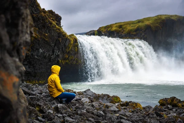아이슬란드의고 다 포스 폭포에서 휴식을 취하고 있는 노란 재킷을 입고 있는 관광객 — 스톡 사진
