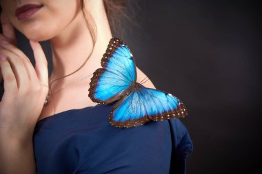 Koyu bir arka planda genç bir kızın omzundaki mavi bir kelebeğin yakın çekimi. Doğanın güzelliği ve kırılganlığı kavramı.