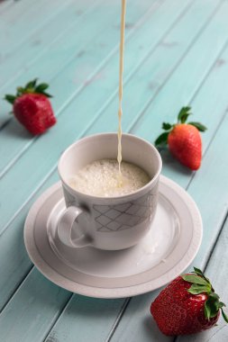 Turkuaz ahşap bir masada beyaz kahve fincanı. Hayattaki çilekler. Süt Kupa 'ya dökülüyor. Bir krema deresi.